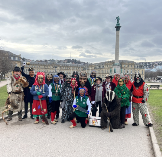 Gruppe von Personen in traditionellen Kostümen und Masken der Kehler Schlammhexe vor dem Jubiläumssäule-Denkmal in Stuttgart.