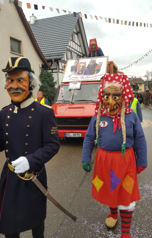 Zwei Personen in rot-weiß gepunkteten Kopftüchern und blauer Kleidung stehen auf der Straße, weitere Personen im Hintergrund bei sonnigem Wetter.